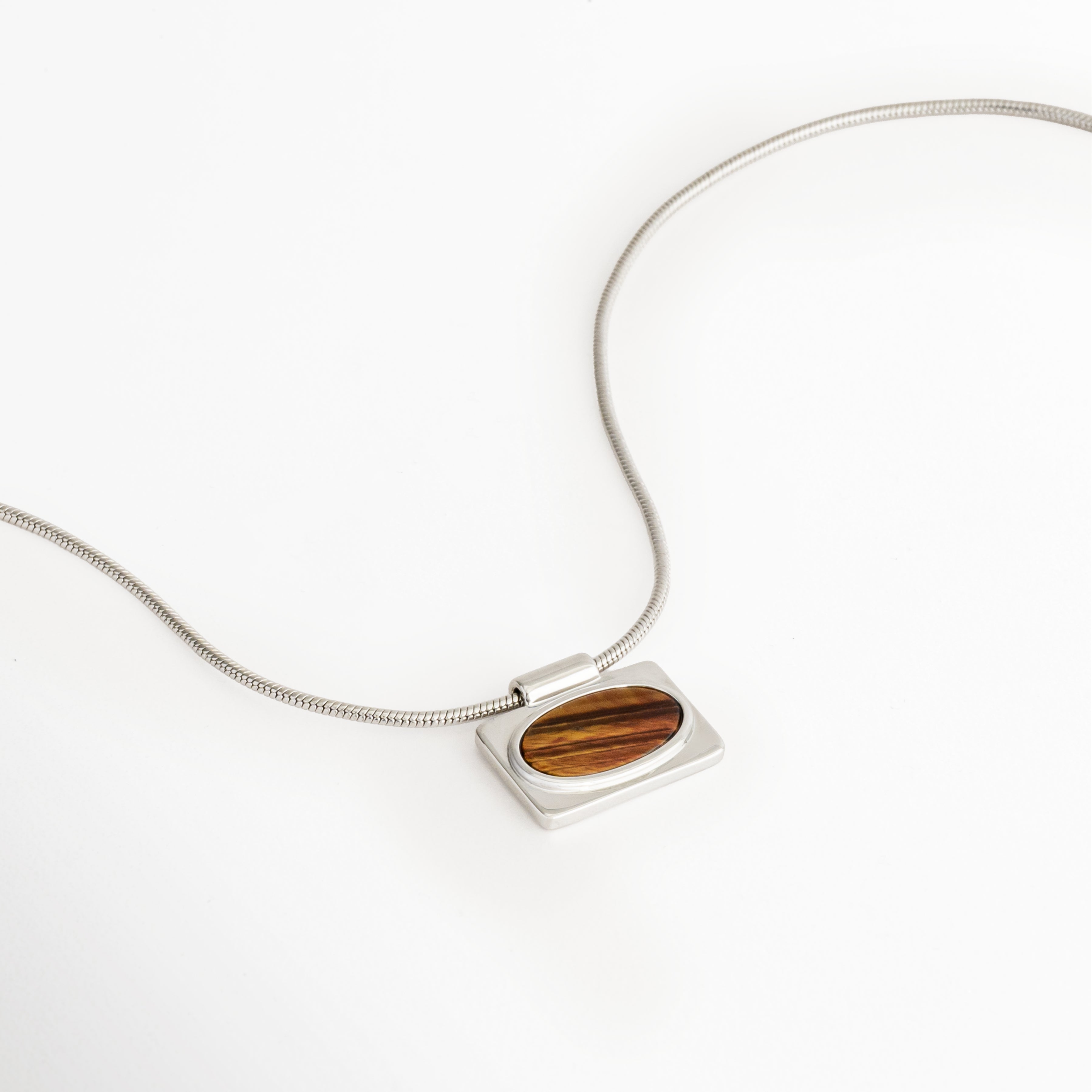 Vista lateral del Collar Tigra mostrando el colgante rectangular con piedra tipo Ojo de Tigre, Collar Tigra en perspectiva que destaca el perfil del dije y la cadena con acabado en Oro Blanco, Vista inclinada del Collar Tigra con piedra Ojo de Tigre y diseño minimalista, Collar Tigra con colgante de piedra natural tipo Ojo de Tigre fotografiado desde un ángulo lateral, Perspectiva del Collar Tigra resaltando el acabado brillante de la cadena y el colgante.
