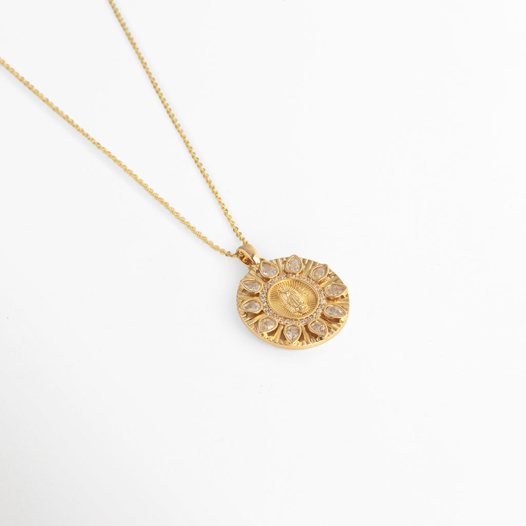 Vista aérea en curva suave del Collar Medalla Virgen Zirconias con el dije circular de 2.3 cm de ancho reposando sobre fondo blanco, Toma extendida de la fina cadena dorada de 45 cm mostrando la soltura del medallón religioso con halo de luz, Muestra de la silueta estilizada y los destellos solares del dije circular en ángulo horizontal, Acercamiento a la caída ligera del collar místico enchapado en oro 14K sobre superficie clara.