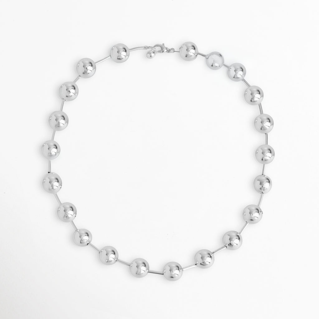 Vista superior en silueta circular del Collar Selene mostrando la distribución de las esferas metálicas enchapadas en oro blanco 24k, Cadena corta plateada dispuesta en círculo que resalta el ritmo uniforme de las esferas de 1.2 cm de diámetro, Plano general aéreo del Collar Selene de 46 cm de longitud extendido de forma circular sobre fondo blanco, Muestra de la estructura simétrica del collar de esferas espaciadas con acabado de alto brillo espejo