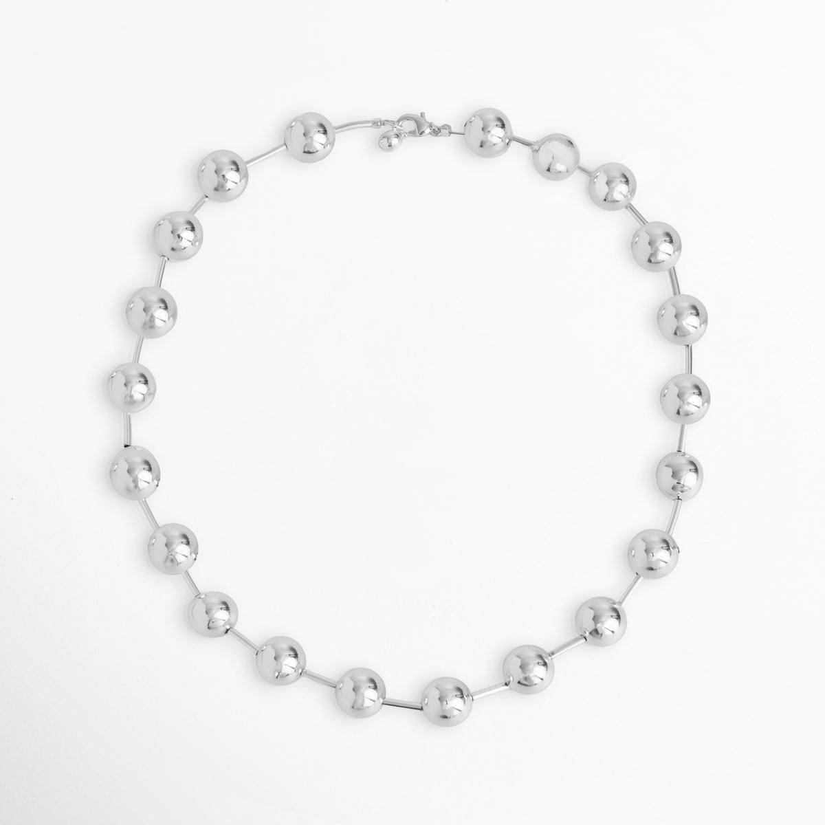 Vista superior en silueta circular del Collar Selene mostrando la distribución de las esferas metálicas enchapadas en oro blanco 24k, Cadena corta plateada dispuesta en círculo que resalta el ritmo uniforme de las esferas de 1.2 cm de diámetro, Plano general aéreo del Collar Selene de 46 cm de longitud extendido de forma circular sobre fondo blanco, Muestra de la estructura simétrica del collar de esferas espaciadas con acabado de alto brillo espejo