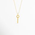Collar Enig de acero inoxidable enchapado en oro 18K con dije de llave orgánica esculpida de 3.5 cm de largo, vista de frente sobre fondo blanco, Vista frontal de collar dorado con cadena serpentina delgada y dije de llave geométrica con cabeza calada, Plano general de collar fino enchapado en oro 18K con dije de llave de estética artesanal y superficie textura suave, Collar Enig destacando la silueta mística de la llave de 1 cm de ancho colgada de una cadena dorada