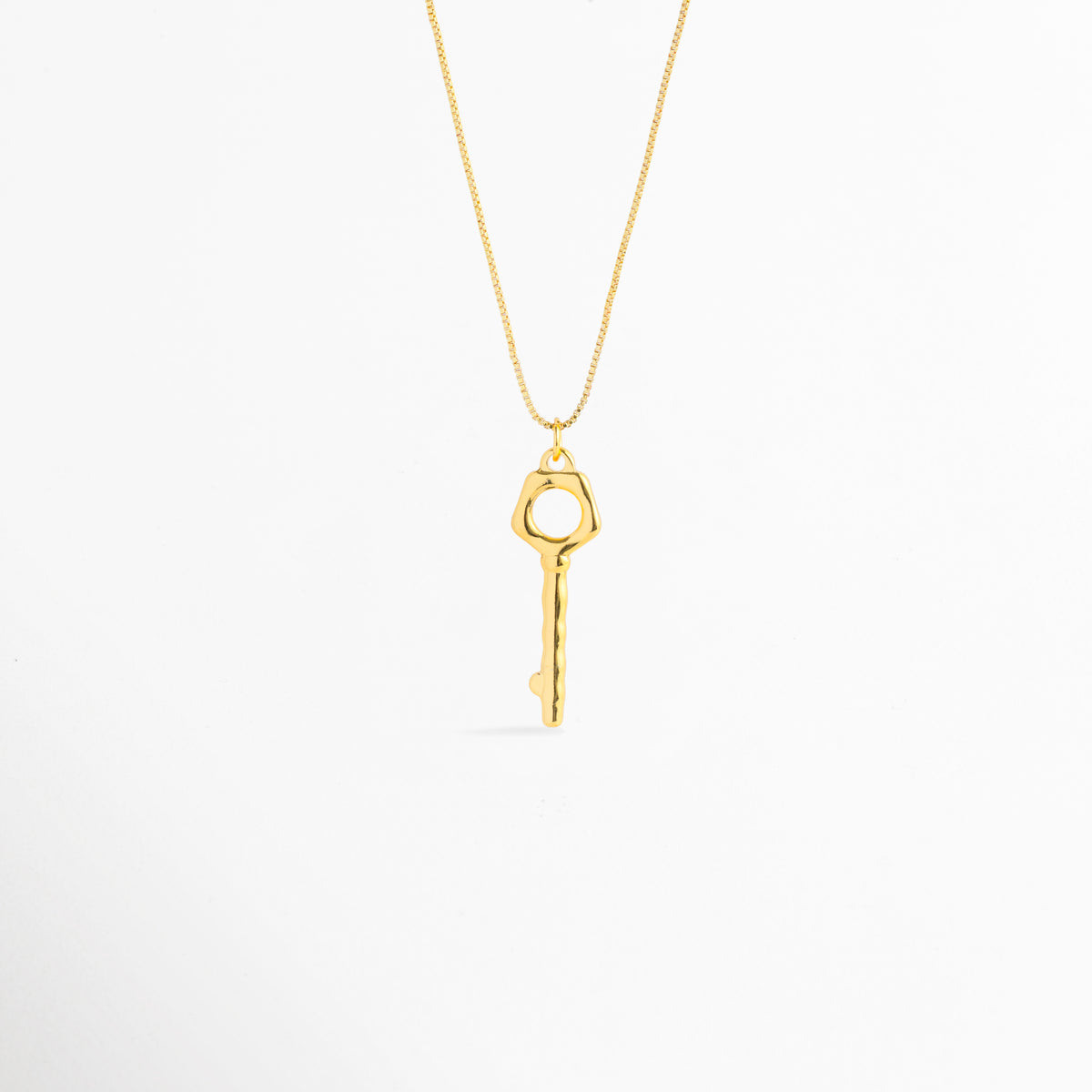 Collar Enig de acero inoxidable enchapado en oro 18K con dije de llave orgánica esculpida de 3.5 cm de largo, vista de frente sobre fondo blanco, Vista frontal de collar dorado con cadena serpentina delgada y dije de llave geométrica con cabeza calada, Plano general de collar fino enchapado en oro 18K con dije de llave de estética artesanal y superficie textura suave, Collar Enig destacando la silueta mística de la llave de 1 cm de ancho colgada de una cadena dorada