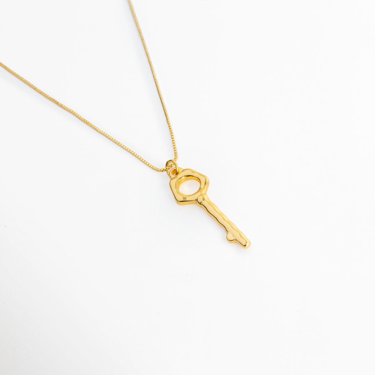 Vista en ángulo del Collar Enig mostrando los contornos orgánicos y el pulido cálido del dije de llave enchapado en oro 18K, Acercamiento en perspectiva al dije de llave dorada con cabeza pentagonal calada suspendido de la cadena lisa, Toma en diagonal que resalta el diseño escultural del dije de 3.5 cm y los reflejos del baño de oro 18K, Muestra de la caída fluida y la silueta arquitectónica del Collar Enig sobre superficie clara.