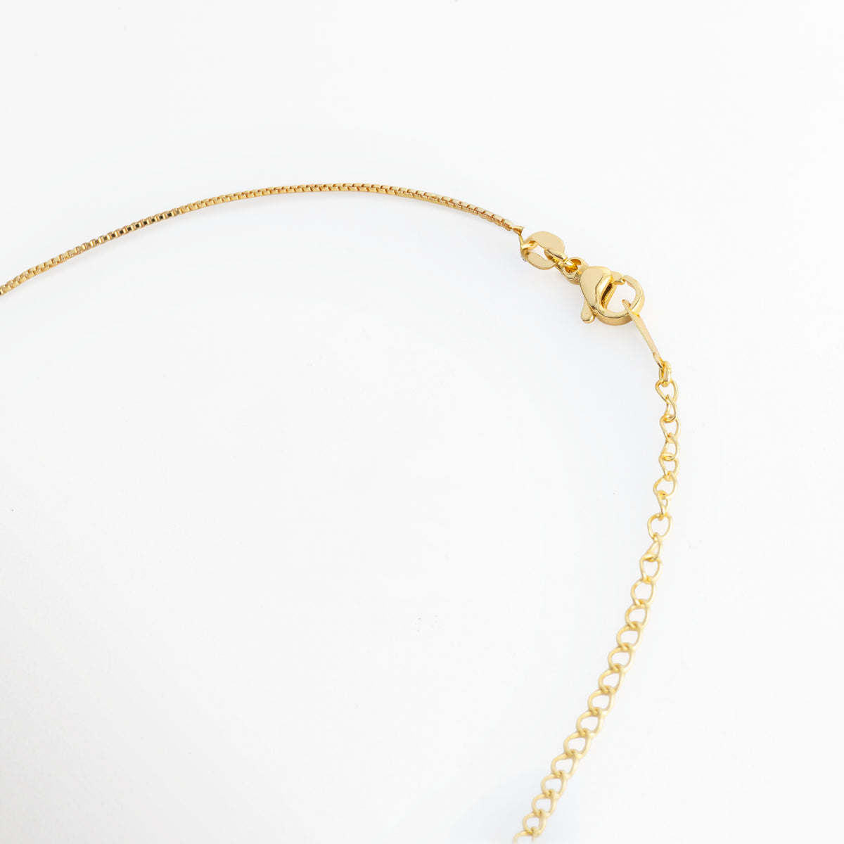 Acercamiento al broche de mosquetón y la cadena de extensión de 6 cm en acero inoxidable enchapado en oro 18K del Collar Enig, Vista macro del sistema de sujeción ajustable y la fina cadena serpentina sobre fondo blanco, Detalle de los eslabones de extensión que permiten adaptar la cadena de 40 cm a 46 cm de longitud total, Muestra del mecanismo de cierre metálico seguro y duradero en tono oro de alto brillo. 
