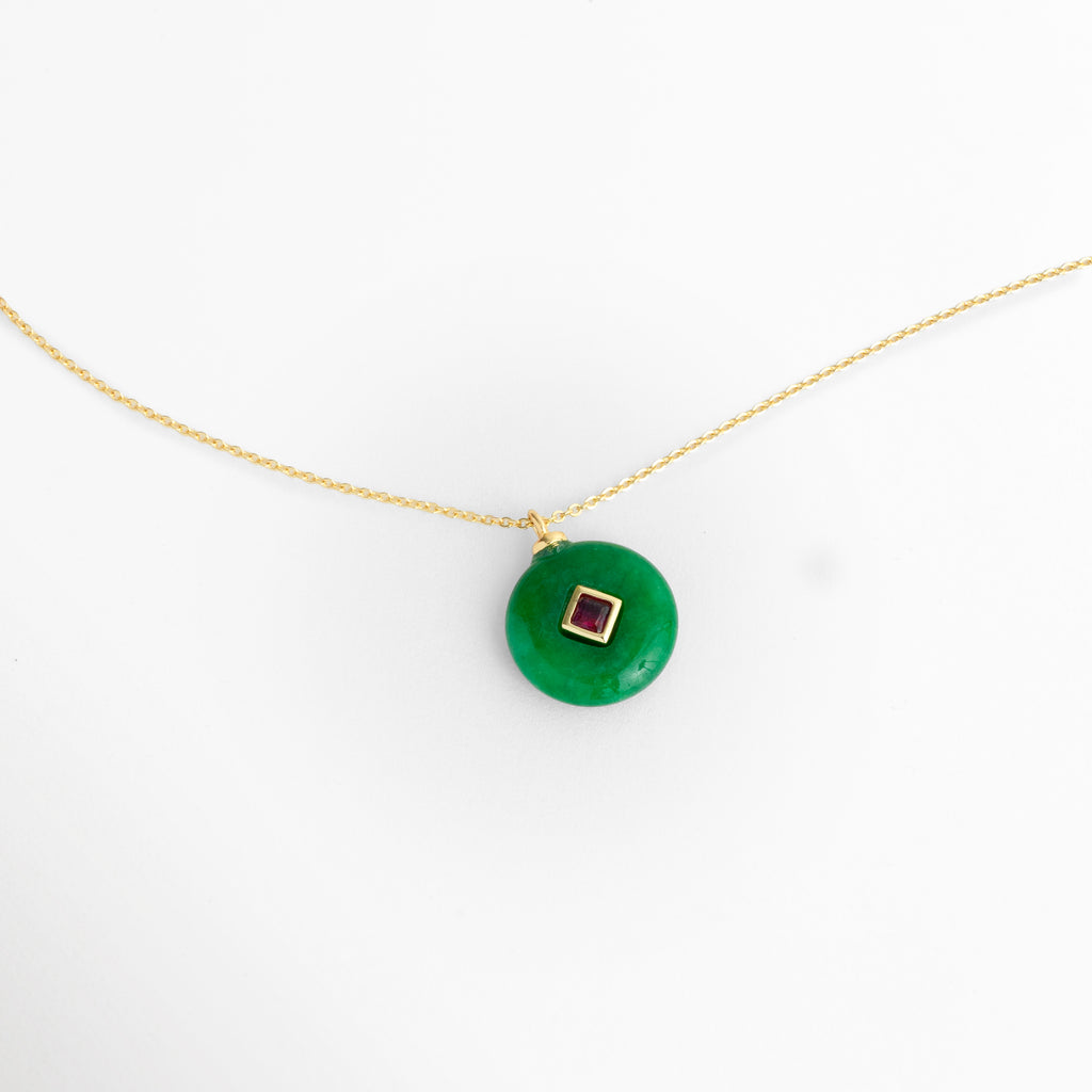 Acercamiento al dije del Collar Ela mostrando la forma de dona o disco plano de la piedra natural verde con zirconia central, Detalle del contraste cromático entre la piedra natural verde y la zirconia en tono fucsia/rubí engastada en rombo, Vista macro del trabajo de enchapado en oro 18K en la argolla de suspensión y el bisel central del dije, Muestra detallada de la superficie lisa y brillante de la piedra natural en forma de medallón circular.