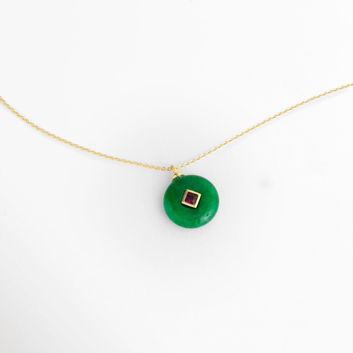 Acercamiento al dije del Collar Ela mostrando la forma de dona o disco plano de la piedra natural verde con zirconia central, Detalle del contraste cromático entre la piedra natural verde y la zirconia en tono fucsia/rubí engastada en rombo, Vista macro del trabajo de enchapado en oro 18K en la argolla de suspensión y el bisel central del dije, Muestra detallada de la superficie lisa y brillante de la piedra natural en forma de medallón circular.