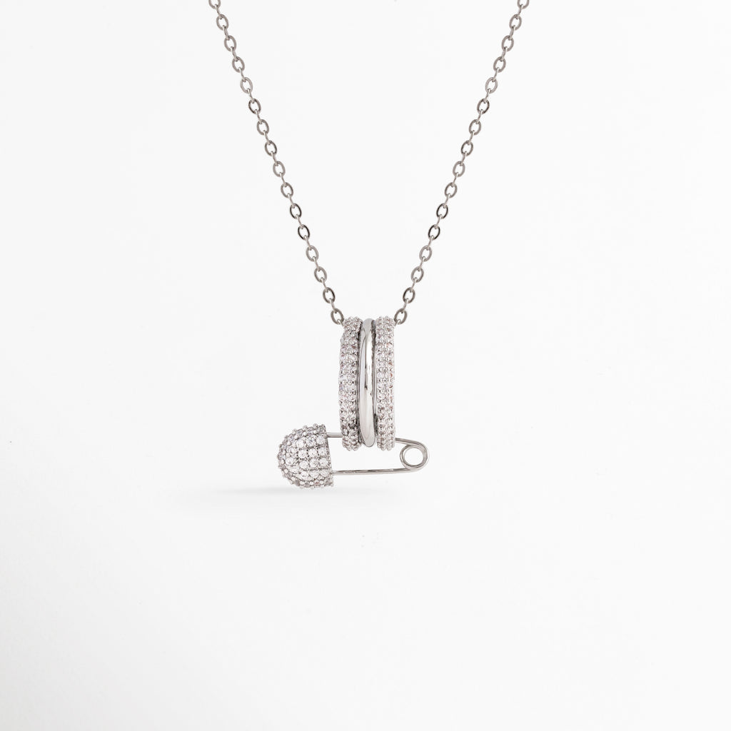 Collar Silver Kravitz con dije en forma de nodriza o imperdible cuajado de circonias y aros entrelazados sobre cadena fina plateada, vista de frente, Vista frontal de collar plateado estilo glam rock con dije de imperdible de 1.7 cm con pavé de circonias transparentes, Plano general de collar plateado con dije de clip de seguridad y aros con pedrería brillante en acabado electrochapado, Collar Silver Kravitz de cobre con dije de imperdible vanguardista de 1.7 x 1.7 cm 