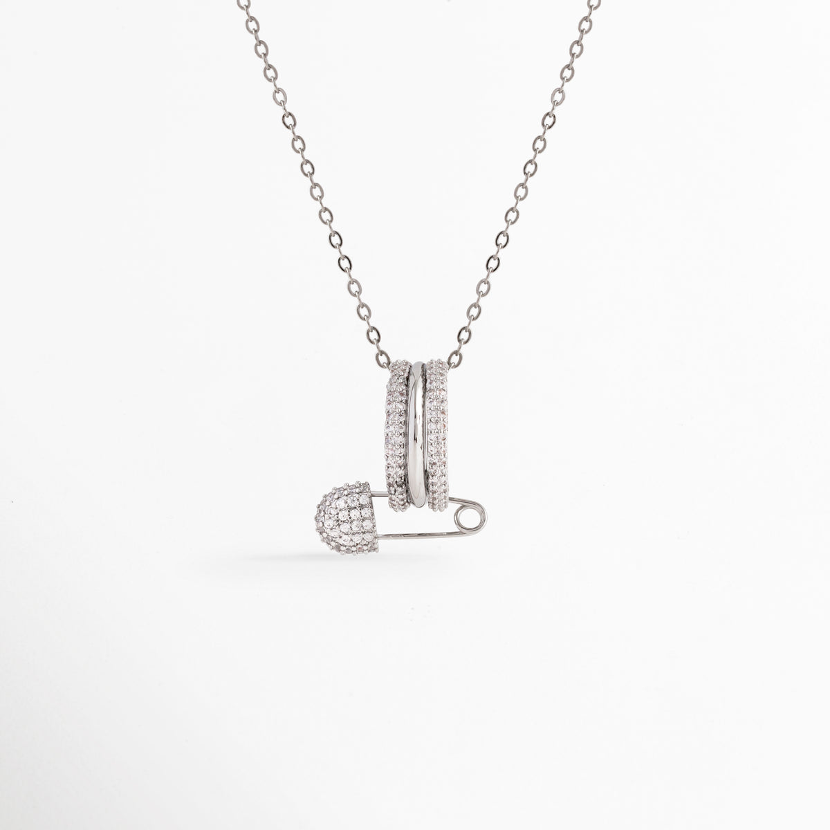Collar Silver Kravitz con dije en forma de nodriza o imperdible cuajado de circonias y aros entrelazados sobre cadena fina plateada, vista de frente, Vista frontal de collar plateado estilo glam rock con dije de imperdible de 1.7 cm con pavé de circonias transparentes, Plano general de collar plateado con dije de clip de seguridad y aros con pedrería brillante en acabado electrochapado, Collar Silver Kravitz de cobre con dije de imperdible vanguardista de 1.7 x 1.7 cm 