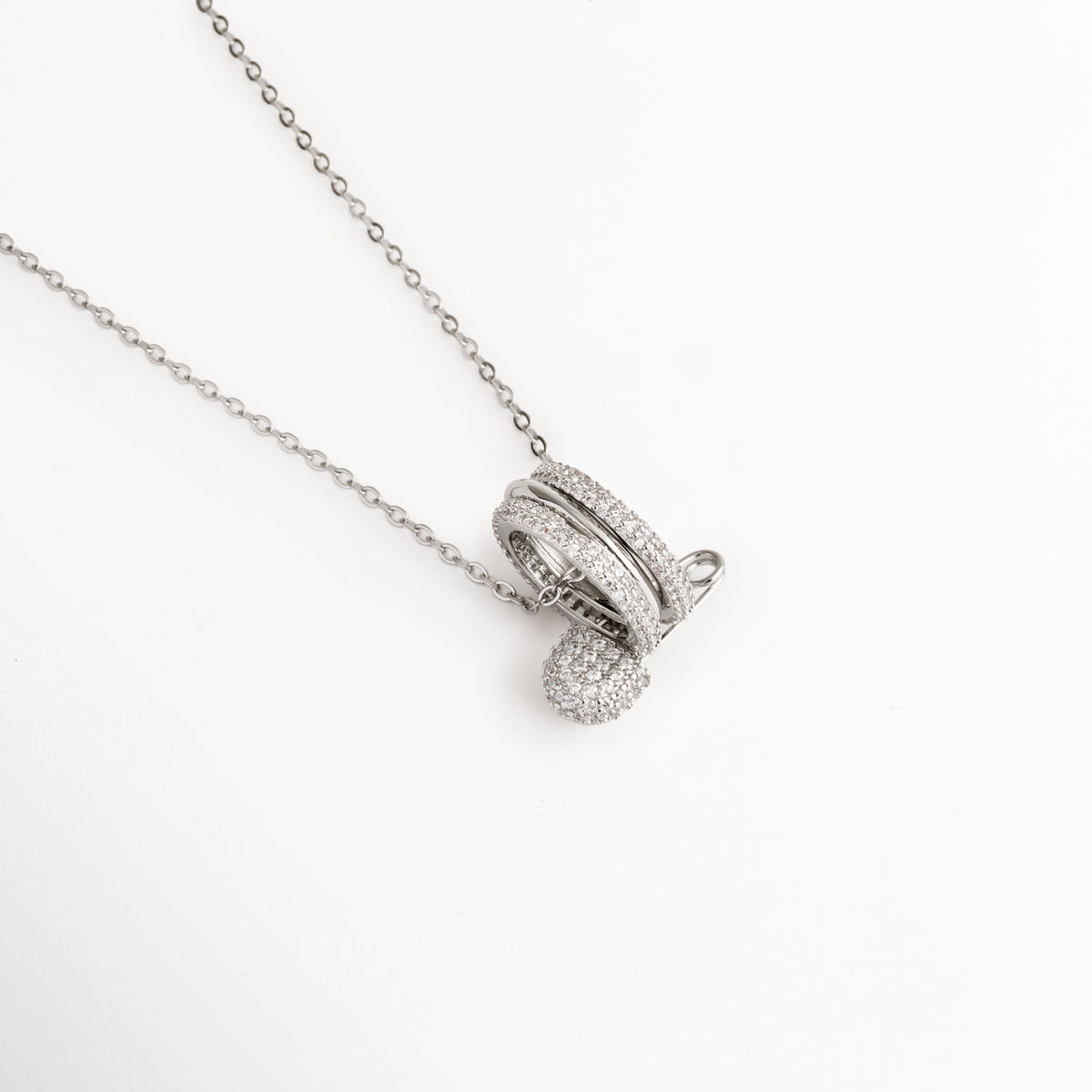 Vista en ángulo del Collar Silver Kravitz mostrando el volumen de los tres aros entrelazados y el imperdible con circonias, Acercamiento en perspectiva al dije de imperdible plateado cuajado de piedras brillantes sobre la cadena fina, Toma en diagonal que resalta el brillo del acabado electrochapado y el relieve de la pedrería cristalina, Muestra de la caída y la proporción del dije de 1.7 cm en la cadena delgada plateada.