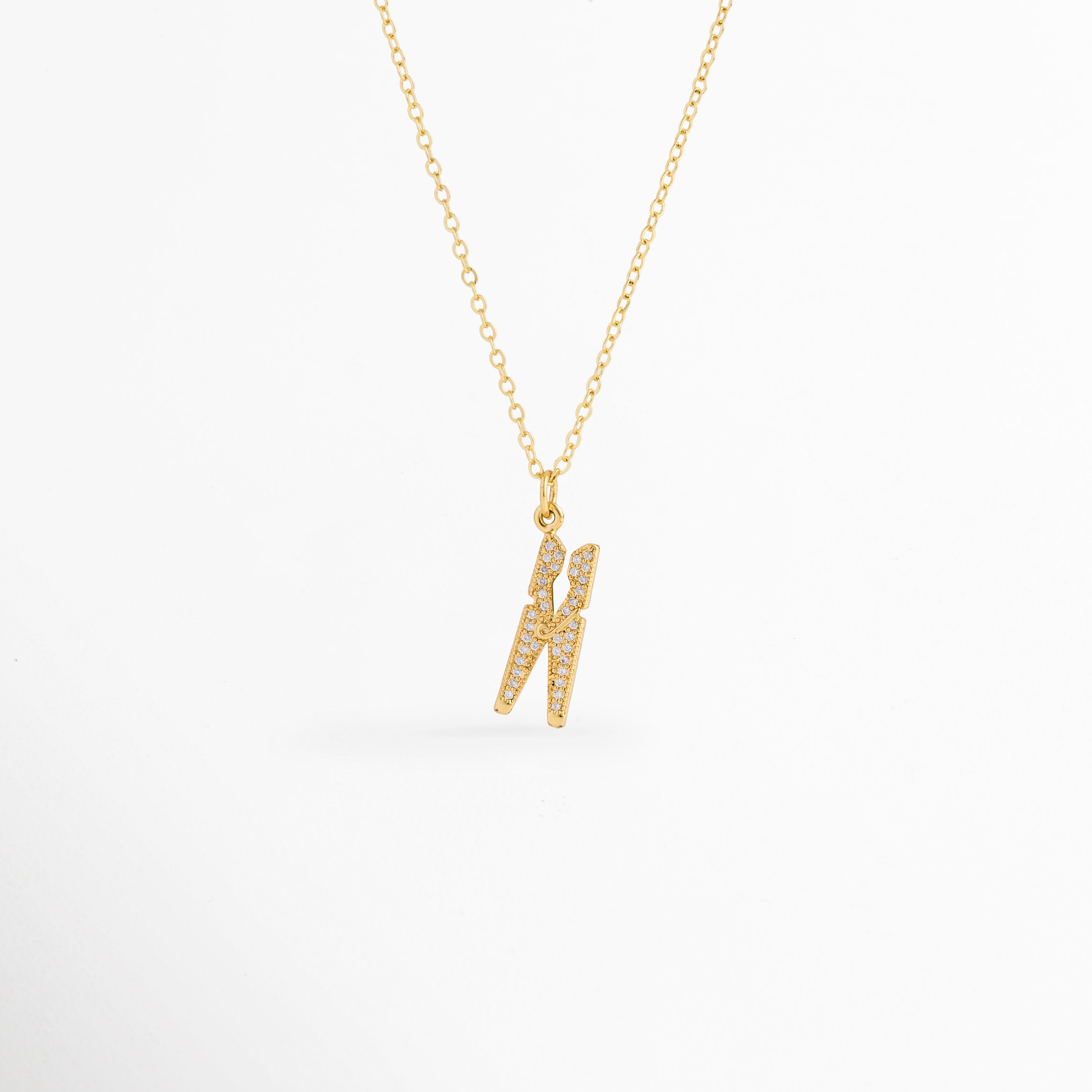 Collar Grapa enchapado en oro 18K con dije de pinza de ropa o gancho cuajado de circonias, vista de frente sobre fondo blanco, Vista frontal de collar dorado con dije de pinza de ropa de 2.5 cm de largo con pavé de circonias brillantes, Plano general de collar con dije de grapa o pinza de ropa vanguardista sobre cadena fina de 40 cm enchapada en oro 18K, Collar Grapa de cobre enchapado en oro 18K mostrando los destellos de las circonias transparentes sobre el dije