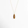 Collar Cuarzo Toffee de cobre enchapado en oro con dije de piedra natural ojo de tigre en forma de gota alargada e incrustación central violeta, vista frontal sobre fondo blanco, Vista de frente de collar dorado con dije de cuarzo tono café toffee de 4 cm de largo y pequeño cristal ovalado incrustado al centro, Plano general de collar fino con dije orgánico en forma de lágrima en piedra natural veteada tono toffee sobre fondo neutro.
