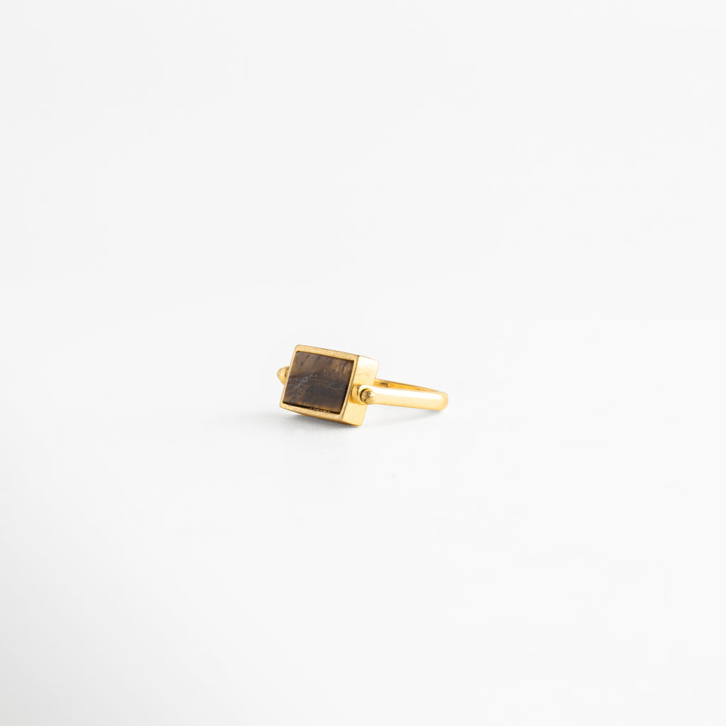 Anillo rectangular de cobre con baño de oro 18K y piedra café natural en ángulo de tres cuartos, Joyas minimalistas: anillo dorado con dije rectangular color tabaco sobre fondo blanco neutro, Detalle de anillo de sello moderno con acabado en oro 18K y piedra semipreciosa rectangular, Accesorio de bisutería fina: anillo con banda delgada dorada y piedra central café veteada, Vista lateral angular de anillo rectangular estilo Malaya Jewelry con recubrimiento de oro premium.