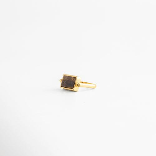 Anillo rectangular de cobre con baño de oro 18K y piedra café natural en ángulo de tres cuartos, Joyas minimalistas: anillo dorado con dije rectangular color tabaco sobre fondo blanco neutro, Detalle de anillo de sello moderno con acabado en oro 18K y piedra semipreciosa rectangular, Accesorio de bisutería fina: anillo con banda delgada dorada y piedra central café veteada, Vista lateral angular de anillo rectangular estilo Malaya Jewelry con recubrimiento de oro premium.