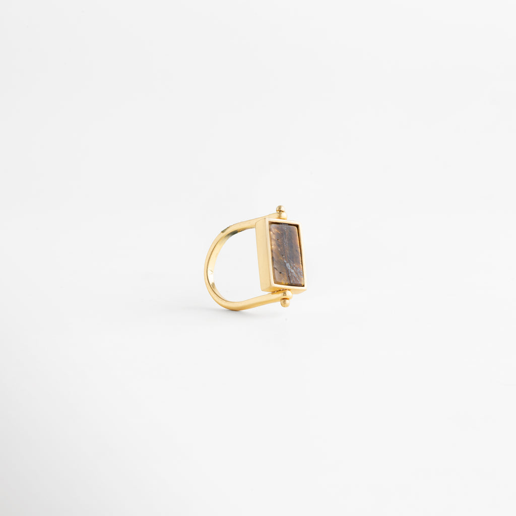 Perfil de anillo dorado mostrando el grosor de la banda de cobre y el soporte del dije, Estructura lateral de anillo rectangular enchapado en oro 18K con diseño de perfil limpio, Vista de costado de anillo minimalista: conexión entre la banda dorada y la piedra café, Perspectiva lateral de joya contemporánea resaltando el pulido del baño de oro de 18 quilates, Foto de perfil de anillo con piedra rectangular que muestra la elevación de la montura dorada.