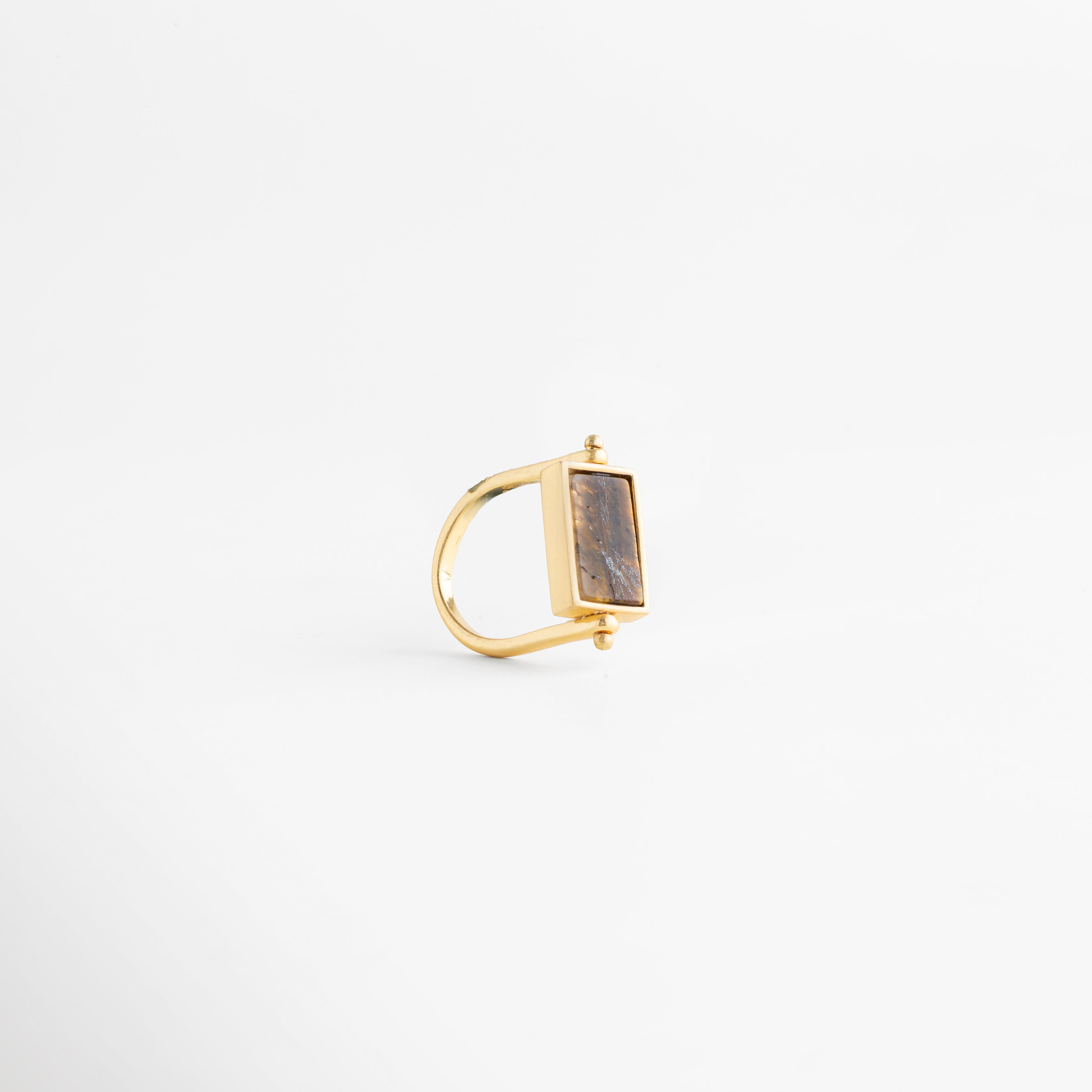 Perfil de anillo dorado mostrando el grosor de la banda de cobre y el soporte del dije, Estructura lateral de anillo rectangular enchapado en oro 18K con diseño de perfil limpio, Vista de costado de anillo minimalista: conexión entre la banda dorada y la piedra café, Perspectiva lateral de joya contemporánea resaltando el pulido del baño de oro de 18 quilates, Foto de perfil de anillo con piedra rectangular que muestra la elevación de la montura dorada.