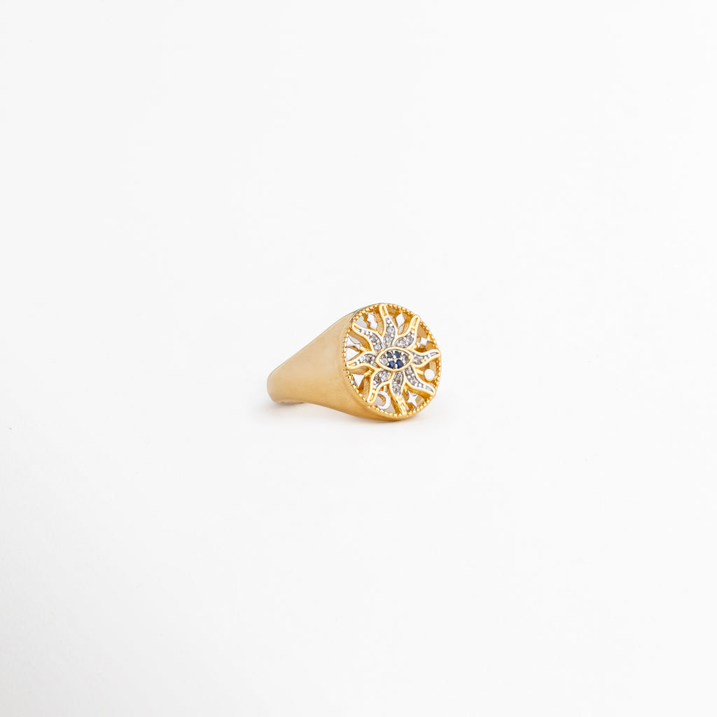 Vista lateral inclinada del anillo Alaric mostrando el relieve de la corona redonda, Detalle del acabado pulido en oro de 14K y la forma cónica del anillo tipo sello, Anillo dorado brillante expuesto en ángulo sobre fondo claro neutro, Enfoque del diseño místico con zirconias capturando la luz de lado, Perspectiva de catálogo del anillo de sol calado chapado en oro.