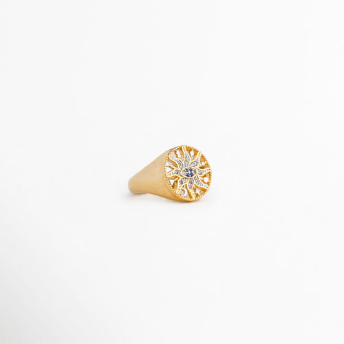 Vista lateral inclinada del anillo Alaric mostrando el relieve de la corona redonda, Detalle del acabado pulido en oro de 14K y la forma cónica del anillo tipo sello, Anillo dorado brillante expuesto en ángulo sobre fondo claro neutro, Enfoque del diseño místico con zirconias capturando la luz de lado, Perspectiva de catálogo del anillo de sol calado chapado en oro.