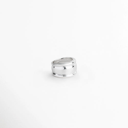 Anillo Tamara plateado en ángulo suave mostrando la abertura interior y el grosor de su silueta cóncava, Vista inclinada de anillo ancho de 1.3 cm en acero inoxidable con esmalte plateado reflectante, Anillo grueso de corte escultórico en perspectiva de tres cuartos sobre fondo claro, Detalle del volumen curvo y los bordes suavemente perfilados de anillo plateado.