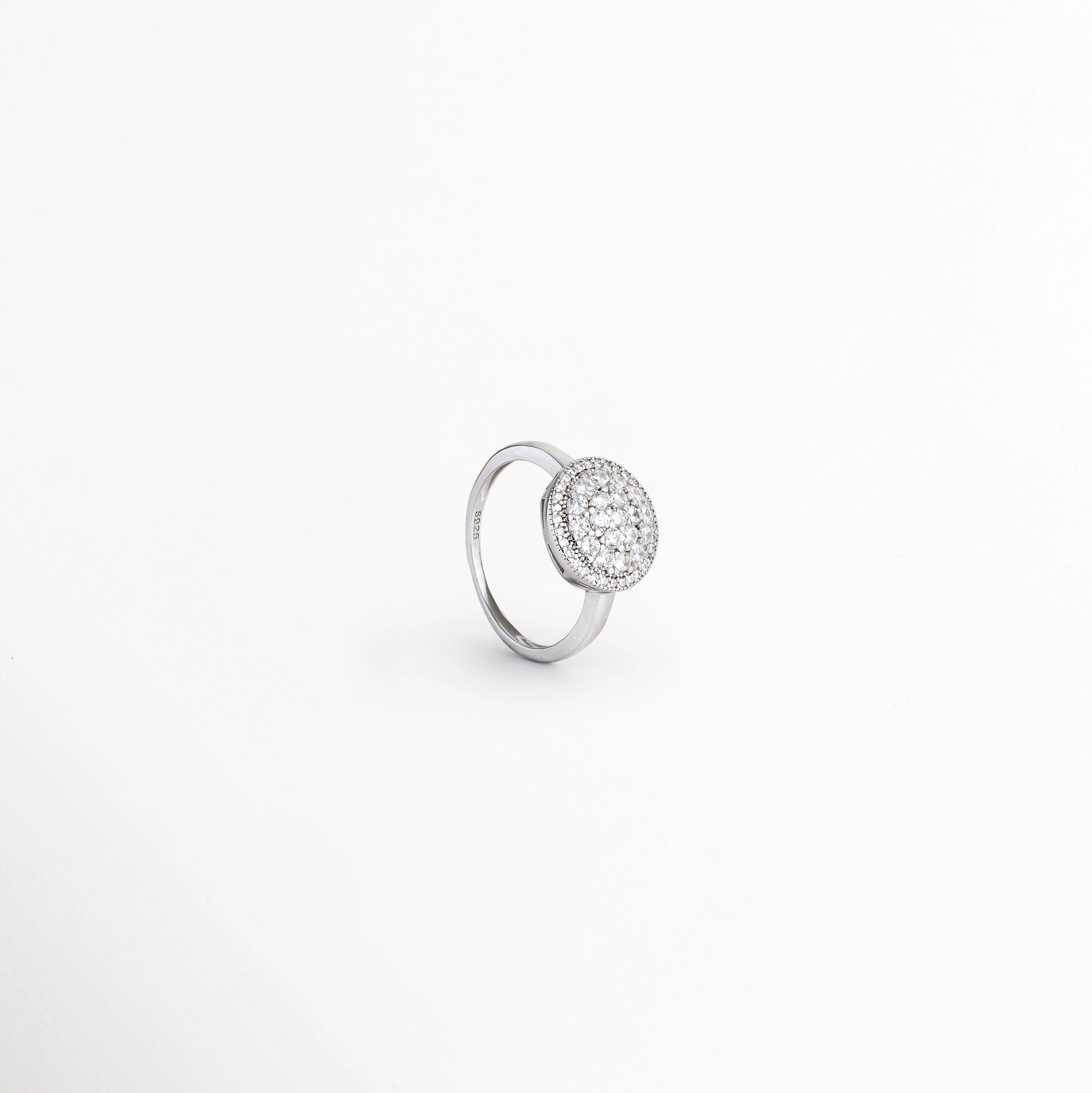 Anillo Tarsila en perspectiva de tres cuartos destacando el resplandor tridimensional de su halo de circonias de 1.8 cm, Vista inclinada de anillo rosetón en Plata Ley 925 mostrando el relieve de sus incrustaciones de pedrería, Anillo de cóctel plateado en ángulo exhibiendo el borde del bisel halo y la superficie engastada, Toma oblicua de anillo elegante de plata 925 con centro redondo lleno de circonias transparentes.