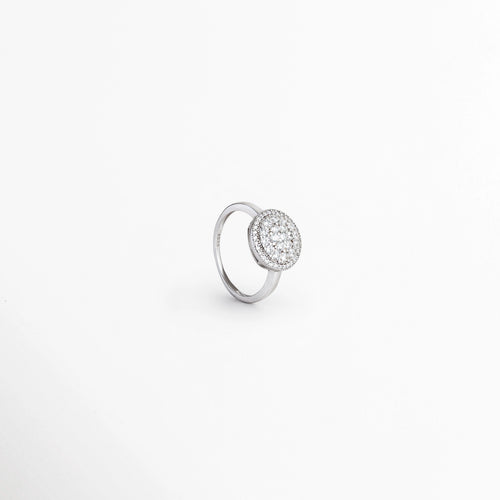 Anillo Tarsila en perspectiva de tres cuartos destacando el resplandor tridimensional de su halo de circonias de 1.8 cm, Vista inclinada de anillo rosetón en Plata Ley 925 mostrando el relieve de sus incrustaciones de pedrería, Anillo de cóctel plateado en ángulo exhibiendo el borde del bisel halo y la superficie engastada, Toma oblicua de anillo elegante de plata 925 con centro redondo lleno de circonias transparentes.