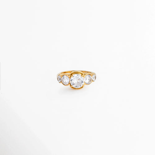 Anillo Fiorela enchapado en oro 18K con incrustación central de zirconia grande flanked por circonias laterales de menor tamaño, vista frontal sobre fondo blanco, Anillo de acero inoxidable dorado con alineación de cinco circonias transparentes de corte brillante en engaste cerrado, Vista de frente de anillo tipo cintillo o compromiso dorado con circonia central destacada sobre superficie clara, Anillo dorado fino de 1 cm de longitud con hileras de circonias en degradé