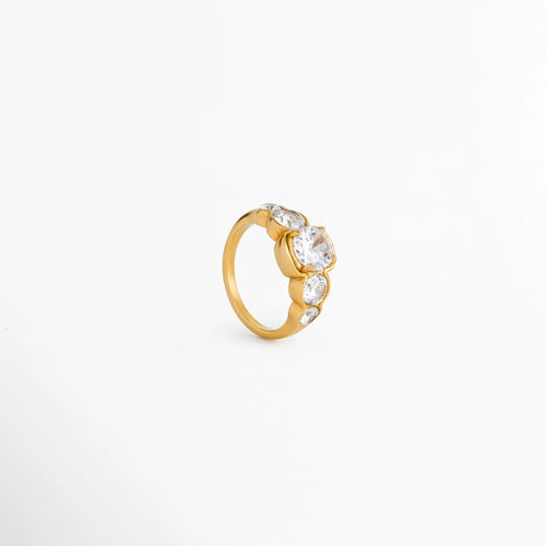 Anillo Fiorela en perspectiva de tres cuartos destacando el destello y facetado de la zirconia central de 1 cm, Vista inclinada de anillo delgado dorado enchapado en oro 18K con fila de circonias deslumbrantes, Anillo estilo cintillo clásico en ángulo de tres cuartos exhibiendo la tridimensionalidad de los engastes, Toma en ángulo de anillo atemporal de acero dorado con incrustaciones cristalinas sobre superficie clara.