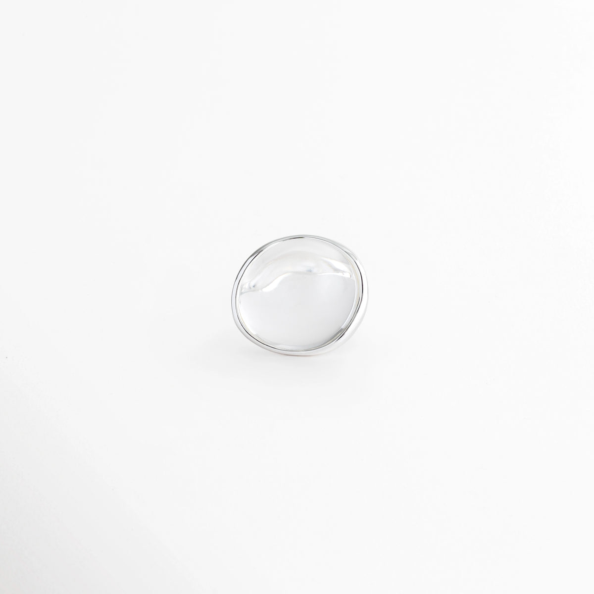 Anillo Palencia enchapado en oro blanco de silueta abombada y ovalada con cabujón de piedra cristalina transparente, vista superior sobre fondo blanco, Vista cenital de anillo cocktail statement plateado con piedra cristalina traslúcida de 2.3 cm de ancho, Plano general de anillo escultural de latón enchapado en oro blanco con domo suave de piedra cristalina transparente, Anillo Palencia destacando la nitidez y pureza de la piedra cristalina engastada en bisel