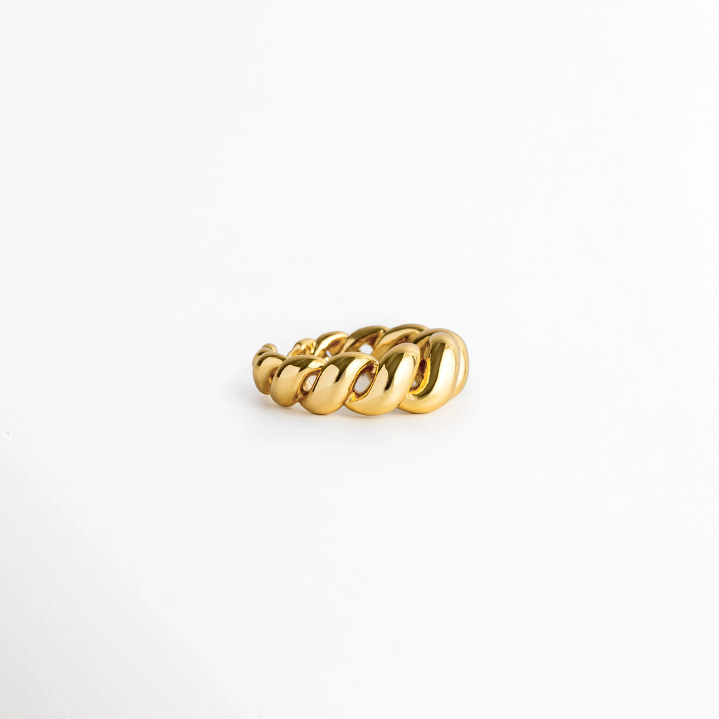 Anillo Olalla Golden visto de perfil acostado sobre fondo blanco, mostrando la curvatura abombada del grabado retorcido, Vista lateral de anillo ajustado enchapado en oro 24K exhibiendo el relieve de sus espirales metálicas, Detalle de perfil de la textura entrelazada tipo cuerda en tono oro brillante, Perspectiva lateral de anillo abierto con relieve en espiral reposando en superficie clara.