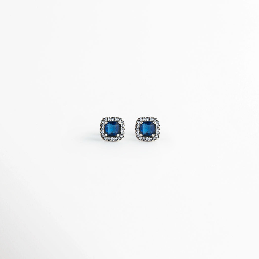 Vista frontal del par de aretes broquel del Set Lunet con circonia azul central de corte cuadrado y halo de micro-pedrería transparente, Acercamiento a los aretes stud cuadrangulares de 0.8 cm de largo enchapados en plata 925 con piedras azul zafiro, Toma de frente de los broqueles de tono plata con gema azul brillante y contorno de circonias cristalinas,  Muestra de la simetría y el contraste elegante entre el azul noche y el acabado metálico plateado.