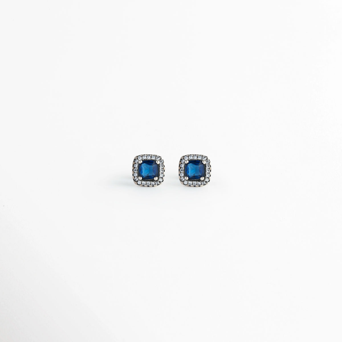 Vista frontal del par de aretes broquel del Set Lunet con circonia azul central de corte cuadrado y halo de micro-pedrería transparente, Acercamiento a los aretes stud cuadrangulares de 0.8 cm de largo enchapados en plata 925 con piedras azul zafiro, Toma de frente de los broqueles de tono plata con gema azul brillante y contorno de circonias cristalinas,  Muestra de la simetría y el contraste elegante entre el azul noche y el acabado metálico plateado.