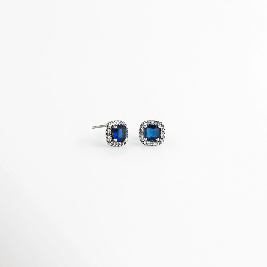 Vista lateral de los aretes del Set Lunet mostrando el poste para oreja perforada y la altura del engaste de la circonia azul, Acercamiento de perfil a la estructura del broquel enchapado en plata 925 y su halo de pedrería fina, Toma en ángulo que resalta la calidad del engaste de la gema central azul de 0.8 cm y la mariposa posterior, Muestra del acabado metálico pulido y la solidez del poste de sujeción para los aretes. 