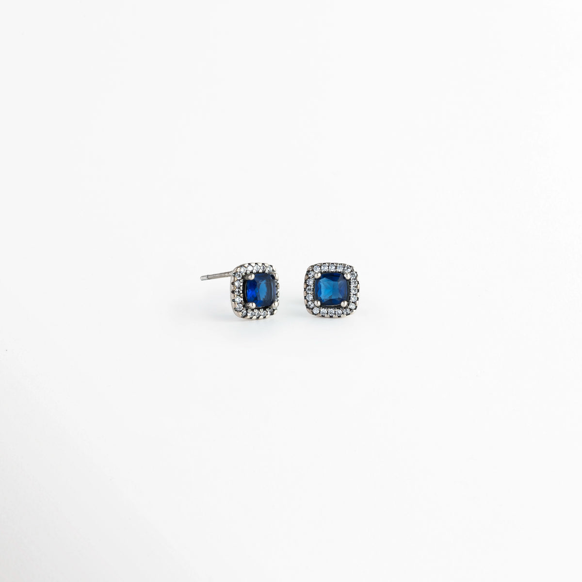 Vista lateral de los aretes del Set Lunet mostrando el poste para oreja perforada y la altura del engaste de la circonia azul, Acercamiento de perfil a la estructura del broquel enchapado en plata 925 y su halo de pedrería fina, Toma en ángulo que resalta la calidad del engaste de la gema central azul de 0.8 cm y la mariposa posterior, Muestra del acabado metálico pulido y la solidez del poste de sujeción para los aretes. 