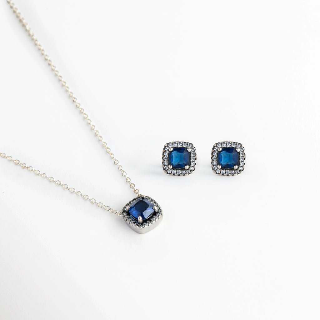 Set Lunet de cobre enchapado en plata 925 compuesto por collar con dije cuadrado y aretes broquel a juego con circonia azul zafiro central y halo transparente, vista de frente sobre fondo blanco, Vista frontal del Set Lunet mostrando el dije de collar de 0.9 cm de ancho y los aretes stud a juego de 0.8 cm en acabado plateado, Plano general de juego de joyería de estilo glam enchapado en plata 925 con circonias centrales color azul profundo y micro-halo de pedrería.