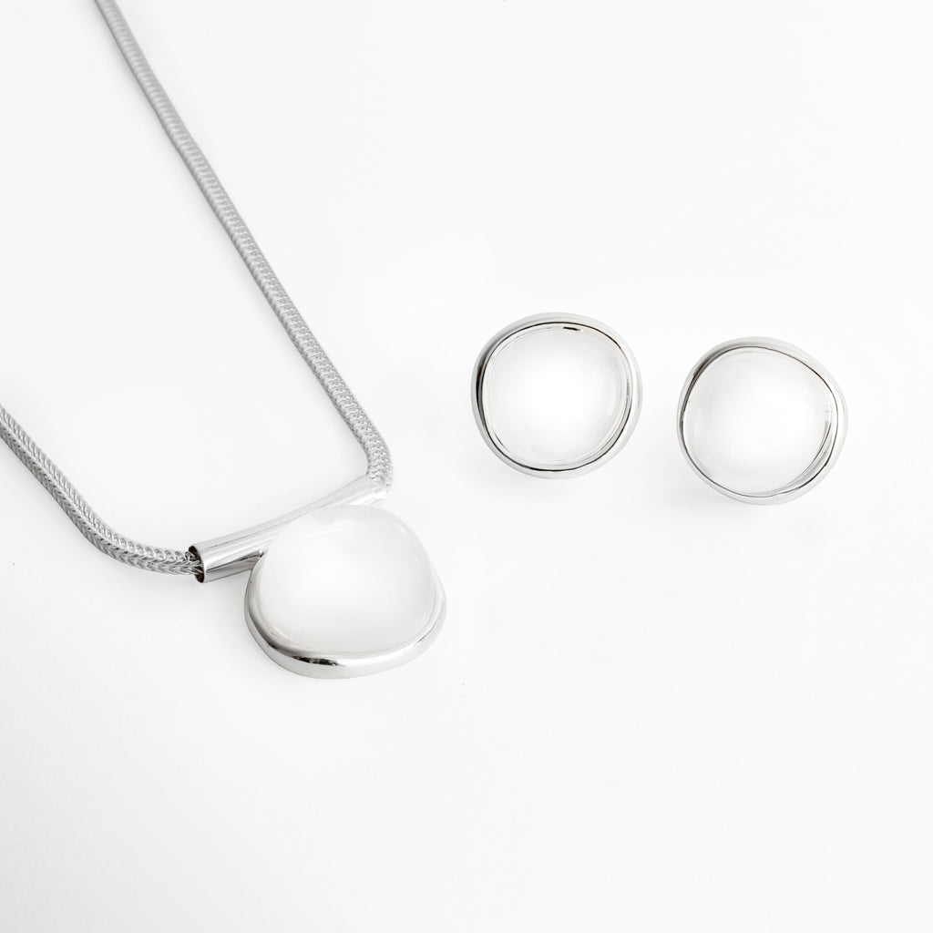 Set Palencia enchapado en oro blanco compuesto por collar con cadena de cola de topo y dije ovalado abombado con piedra cristalina traslúcida junto a aretes broquel a juego, vista superior sobre fondo blanco, Vista frontal del Set Palencia mostrando el dije del collar de 2.7 cm de ancho y los aretes circulares abombados de 2.2 cm en acabado plateado, Plano general de juego de joyería de latón enchapado en oro blanco con piedras cristalinas transparentes de estilo escultural
