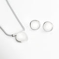 Set Palencia enchapado en oro blanco compuesto por collar con cadena de cola de topo y dije ovalado abombado con piedra cristalina traslúcida junto a aretes broquel a juego, vista superior sobre fondo blanco, Vista frontal del Set Palencia mostrando el dije del collar de 2.7 cm de ancho y los aretes circulares abombados de 2.2 cm en acabado plateado, Plano general de juego de joyería de latón enchapado en oro blanco con piedras cristalinas transparentes de estilo escultural