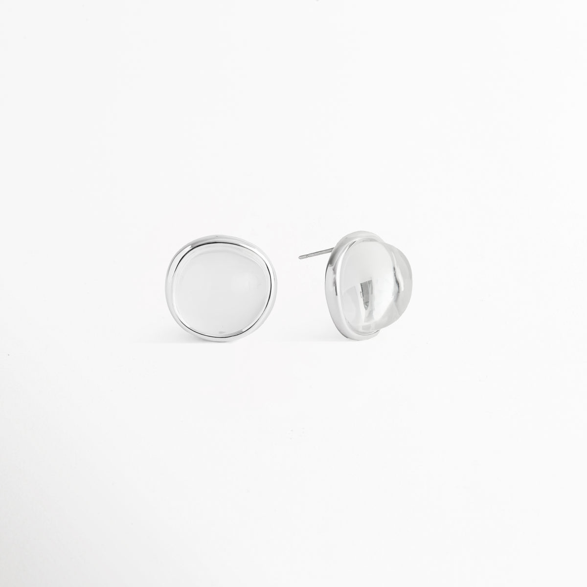 Vista de perfil del par de aretes broquel del Set Palencia mostrando el poste para oreja perforada y la curvatura del domo cristalino, Acercamiento lateral a los aretes abombados de 2.2 cm enchapados en oro blanco con bisel metálico pulido, Toma en ángulo de los aretes de piedra cristalina traslúcida sobre superficie clara, Muestra de la elevación y volumen tridimensional de los broqueles del Set Palencia. 