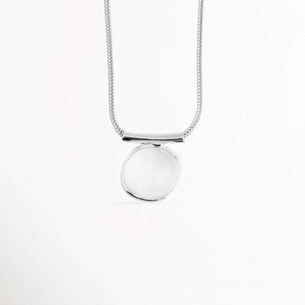 Vista centrada del dije ovalado abombado de 2.7 cm del Collar Palencia colgado de una cadena lisa estilo cola de topo enchapada en oro blanco, Acercamiento al dije de piedra cristalina transparente con pasador metálico horizontal pulido, Toma de frente del collar mostrando la simetría y la claridad de la gema cristalina traslúcida, Muestra del brillo pulido espejo de la montura de latón enchapado sobre fondo blanco. 