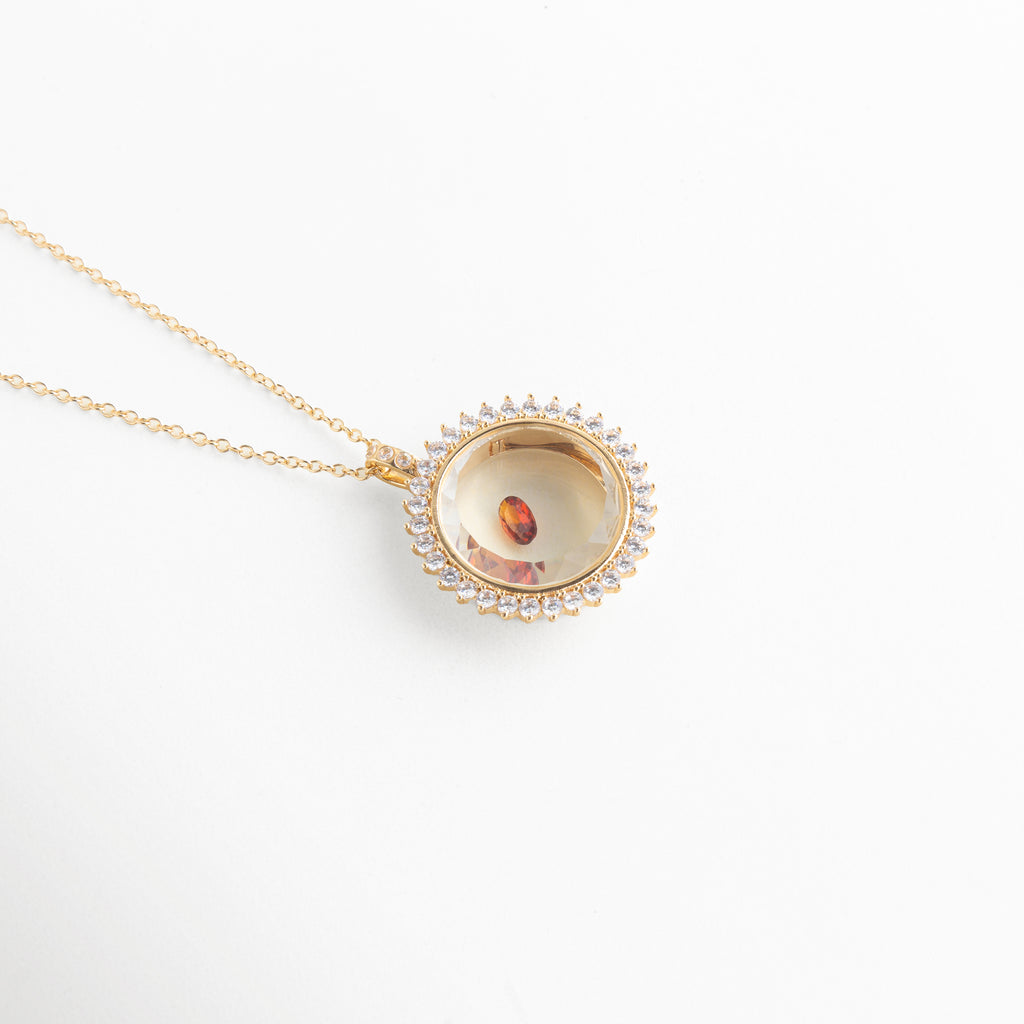 Collar dorado con dije circular y piedra de cristal color ámbar, Dije elegante con cristal central en tonos cálidos y acabado brillante, Joyería fina con piedra artificial de cristal en diseño delicado, Collar minimalista con cristal pequeño de color rojo anaranjado, Accesorio de bisutería con gema de cristal brillante en diseño moderno