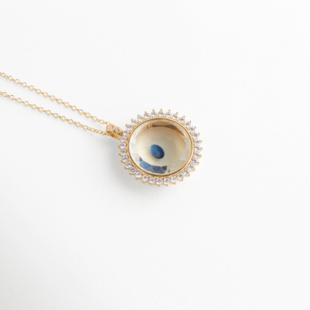 Collar dorado con dije circular y piedra de cristal azul central, Dije elegante con cristal azul brillante en diseño tipo ojo, Joyería fina con piedra azul de cristal en accesorio minimalista, Collar delicado con cristal azul pequeño en acabado brillante, Accesorio de bisutería con gema azul en diseño moderno y fino