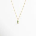 Charm Niño Verde colgado de una fina cadena dorada con dije estilizado en forma de niño con circonia rectangular verde y cabeza de circonia transparente, Collar dorado con dije de figura de niño de 1.09 x 0.8 cm enchapado en oro 18K con gema verde esmeralda en el torso, Vista de frente de dije de silueta de niño en tono oro con vestido/cuerpo de circonia verde baguette sobre fondo blanco, Ejemplo de estilismo del Charm Niño Verde integrado como punto de color