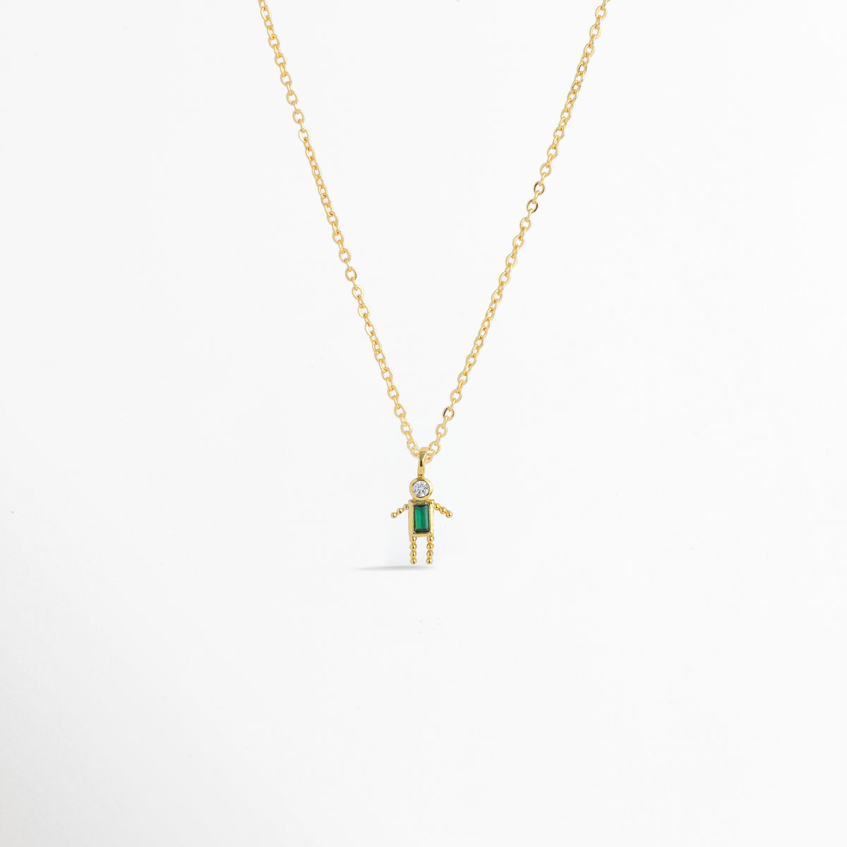 Charm Niño Verde colgado de una fina cadena dorada con dije estilizado en forma de niño con circonia rectangular verde y cabeza de circonia transparente, Collar dorado con dije de figura de niño de 1.09 x 0.8 cm enchapado en oro 18K con gema verde esmeralda en el torso, Vista de frente de dije de silueta de niño en tono oro con vestido/cuerpo de circonia verde baguette sobre fondo blanco, Ejemplo de estilismo del Charm Niño Verde integrado como punto de color