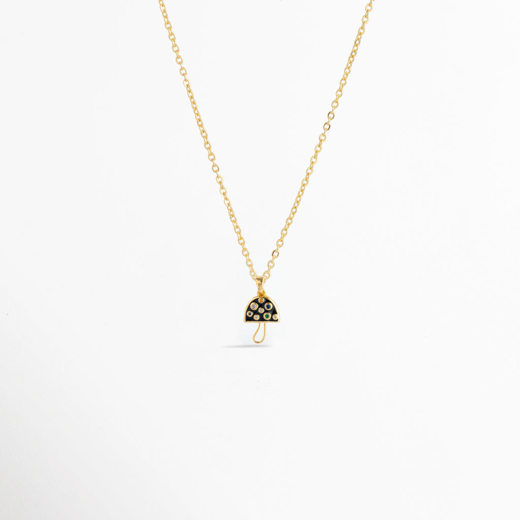 Charm Hongo Negro montado como dije central en una fina cadena dorada sobre fondo blanco, Vista de collar con dije pequeño de hongo en esmalte negro y pedrería multicolor de 2.1 cm enchapado en oro 18K, Perspectiva de gargantilla delicada con dije colgante en forma de seta negra y dorada con circonias, Muestra de escala del charm de hongo negro incrustado con gemas en una cadena delgada enchapada en oro.