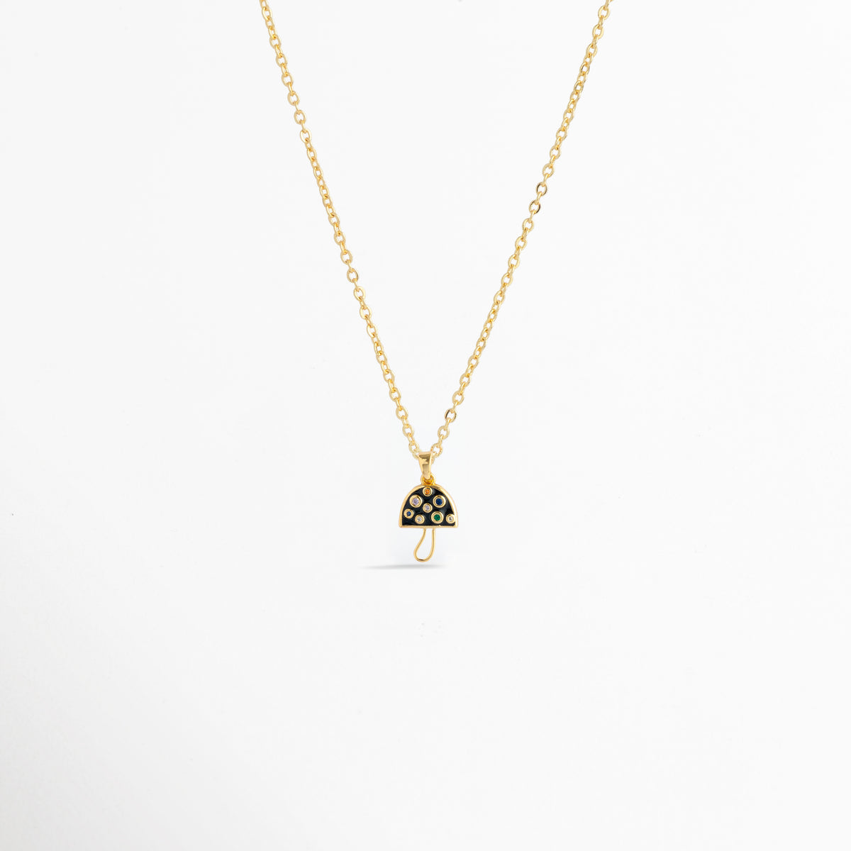 Charm Hongo Negro montado como dije central en una fina cadena dorada sobre fondo blanco, Vista de collar con dije pequeño de hongo en esmalte negro y pedrería multicolor de 2.1 cm enchapado en oro 18K, Perspectiva de gargantilla delicada con dije colgante en forma de seta negra y dorada con circonias, Muestra de escala del charm de hongo negro incrustado con gemas en una cadena delgada enchapada en oro.