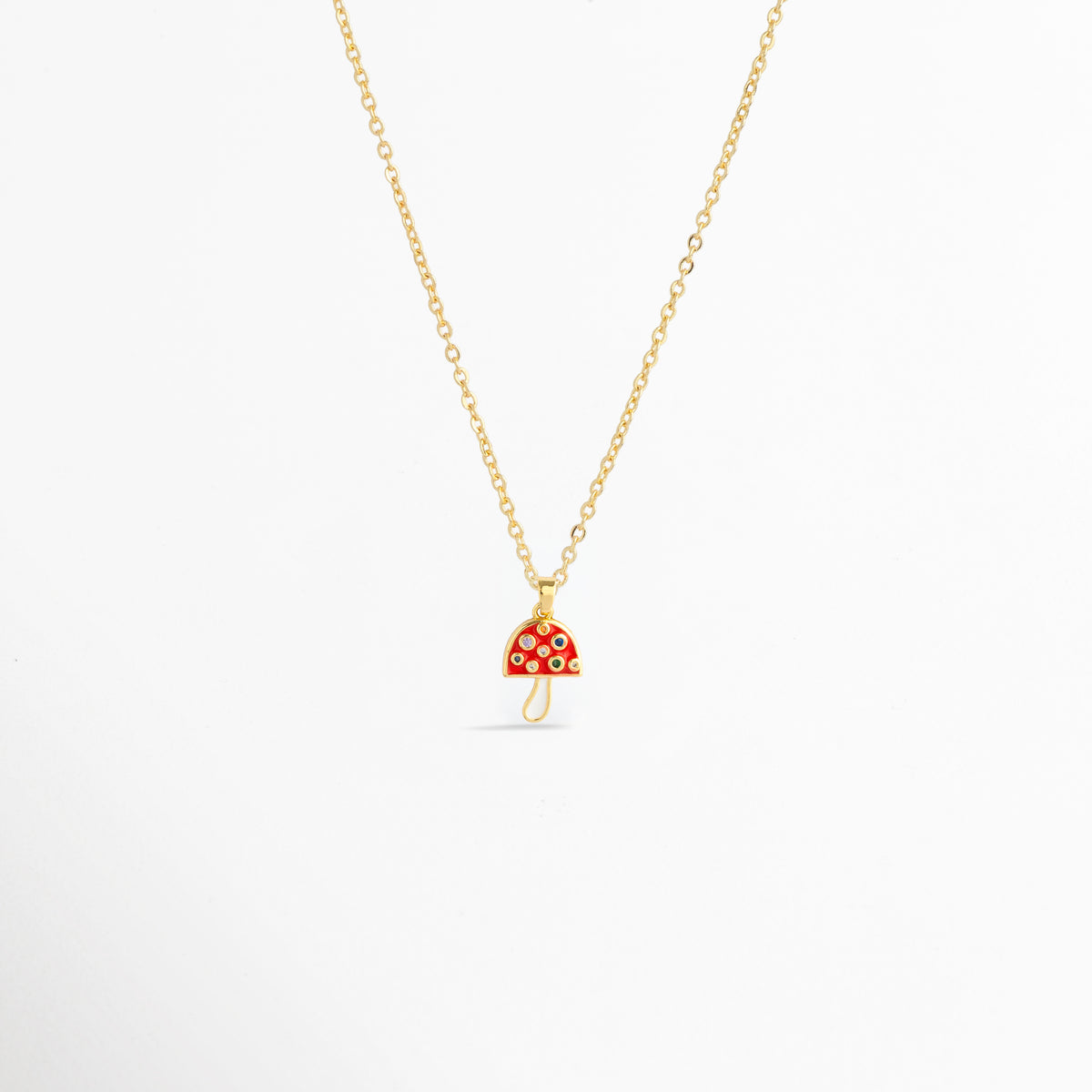 Charm Hongo Rojo montado como dije central en una fina cadena dorada sobre fondo blanco, Vista de collar con dije pequeño de hongo en esmalte rojo y pedrería multicolor de 2.1 cm enchapado en oro 18K, Perspectiva de gargantilla delicada con dije colgante en forma de seta roja y dorada con circonias, Muestra de escala del charm de hongo rojo incrustado con gemas en una cadena delgada enchapada en oro.
