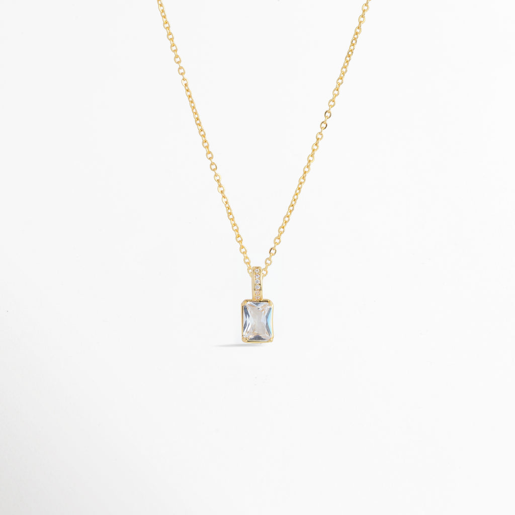 Charm Shimmer colocado como colgante central en una delicada cadena dorada, vista frontal en plano general, Collar dorado con dije rectangular de circonia transparente y corte brillante de estilo atemporal, Vista de colgante en forma de gema rectangular sobre cadena fina dorada enchapada sobre fondo blanco, Ejemplo de estilismo del Charm Shimmer usado como dije central para gargantilla o collar elegante.