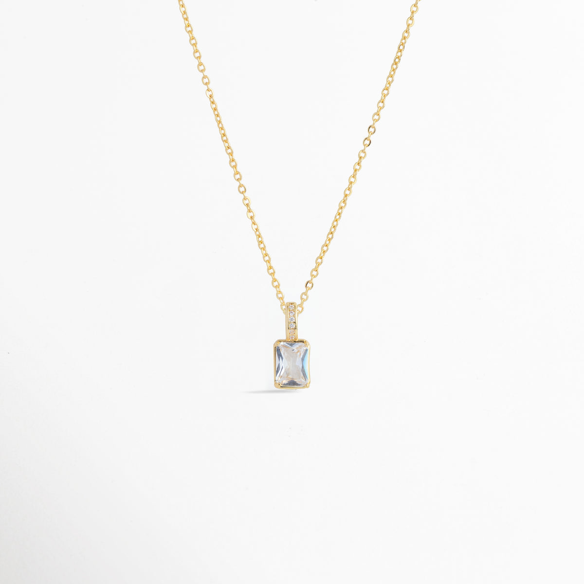 Charm Shimmer colocado como colgante central en una delicada cadena dorada, vista frontal en plano general, Collar dorado con dije rectangular de circonia transparente y corte brillante de estilo atemporal, Vista de colgante en forma de gema rectangular sobre cadena fina dorada enchapada sobre fondo blanco, Ejemplo de estilismo del Charm Shimmer usado como dije central para gargantilla o collar elegante.