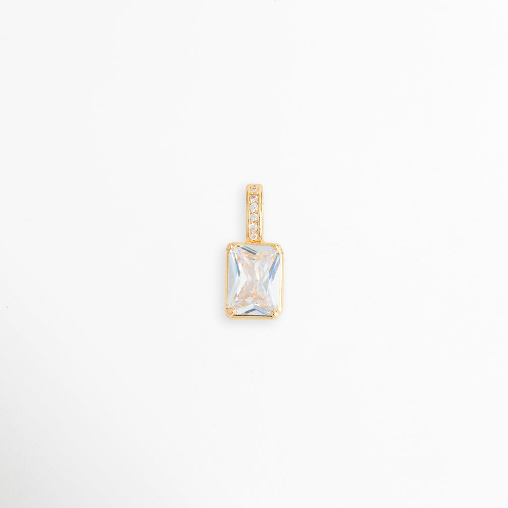 Charm Shimmer con gran circonia transparente de corte esmeralda y reasa vertical con micro-pavé, vista frontal sobre fondo blanco, Dije rectangular de 1.1 x 0.7 cm con cristal de circonia en engaste de bisel sobre metal dorado galvanizado, Vista de frente de dije charm de cobre dorado con gema baguette/esmeralda y asa decorada con pedrería, Charm de circonia transparente de corte rectangular con baño galvanizado brillante sobre superficie neutra.