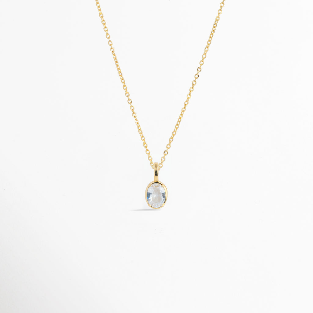 Charm Simple Glow colocado como dije solitario central en una delicada cadena dorada, vista frontal en plano general, Collar dorado estilizado con dije ovalado de circonia transparente de inspiración clásica, Vista de colgante en forma de gota u óvalo brillante sobre cadena fina enchapada sobre fondo blanco, Ejemplo de uso del Charm Simple Glow como punto de luz en una gargantilla dorada minimalista.