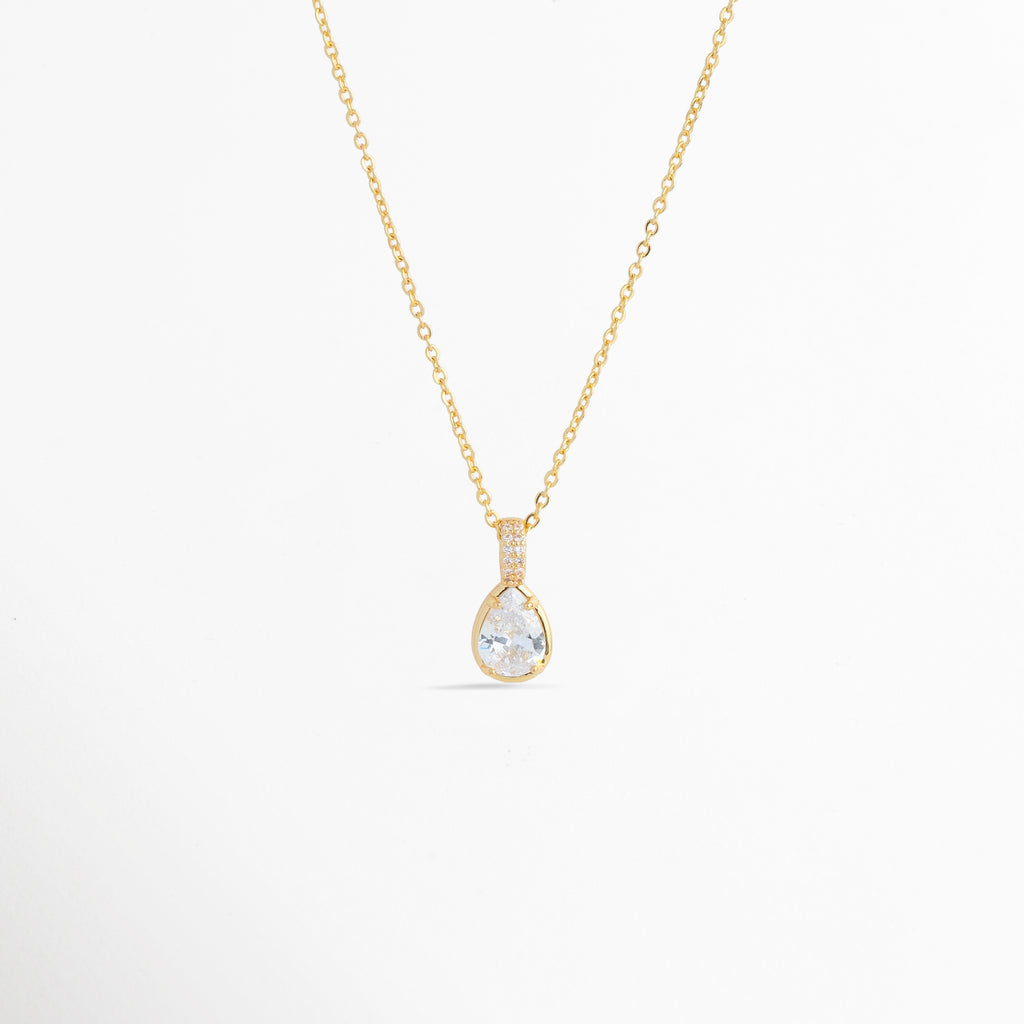 Charm Glow colocado como dije colgante en forma de gota en una delicada cadena dorada, vista frontal en plano general, Collar dorado sofisticado con dije en forma de lágrima de circonia transparente de inspiración romántica, Vista de colgante en forma de gota de luz sobre cadena fina enchapada sobre fondo blanco, Ejemplo de estilismo del Charm Glow usado como dije central para gargantilla o collar elegante.