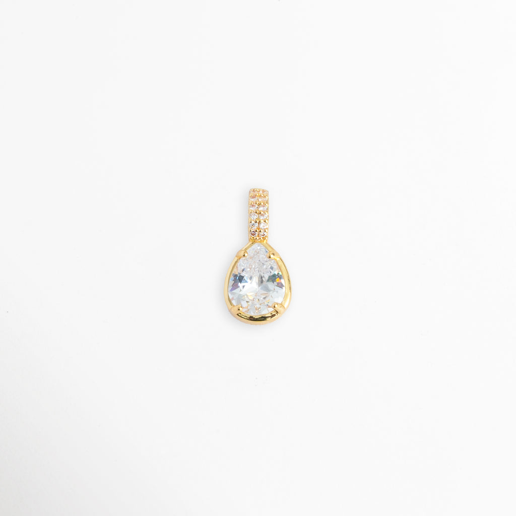Charm Glow con gran circonia transparente en forma de gota o lágrima y reasa vertical con micro-pavé, vista frontal sobre fondo blanco, Dije en forma de lágrima de 1.1 x 0.7 cm con gema de circonia brillante y engaste de bisel sobre metal dorado galvanizado, Vista de frente de dije charm de cobre dorado con gema de talla pera y asa decorada con pedrería incrustada, Charm de circonia transparente de corte gota con recubrimiento galvanizado brillante.